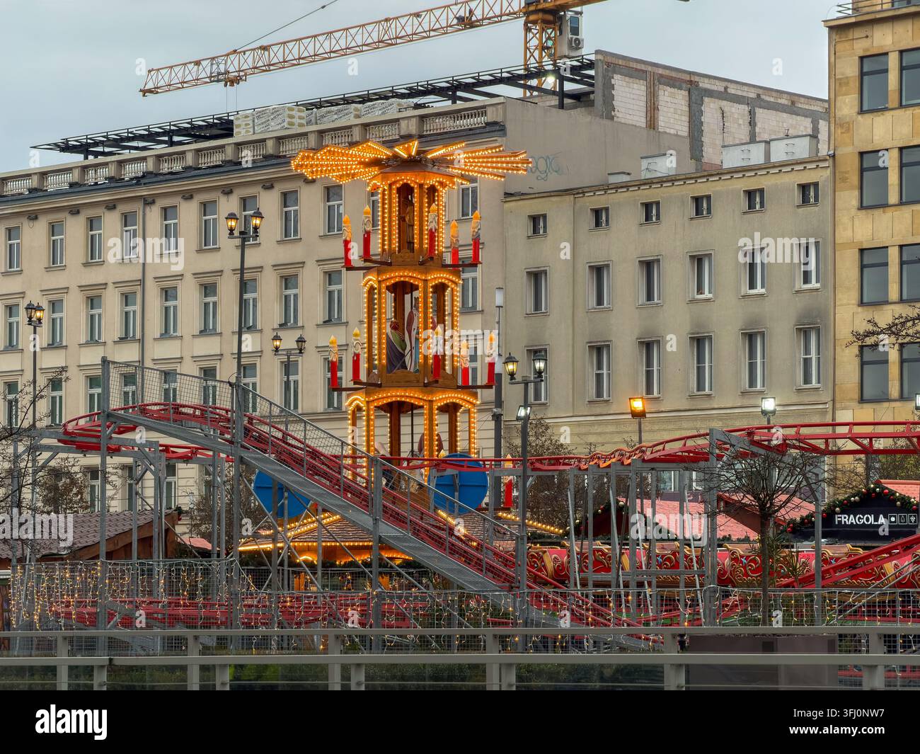 Poznań, Pologne – 23 décembre 2025 : Une tour de manège décorée et lumineuse trône au centre d'une scène de marché saisonnier mettant en vedette une piste de montagne russe en métal sous un c Banque D'Images
