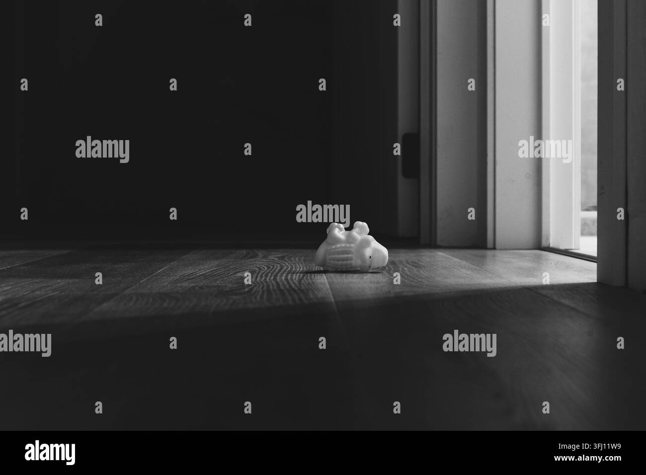 Photographie en noir et blanc à faible angle d'un petit jouet abandonné sur un sol en bois sombre près d'une porte ouverte laissant entrer une lumière vive. Mo mélancolique Banque D'Images