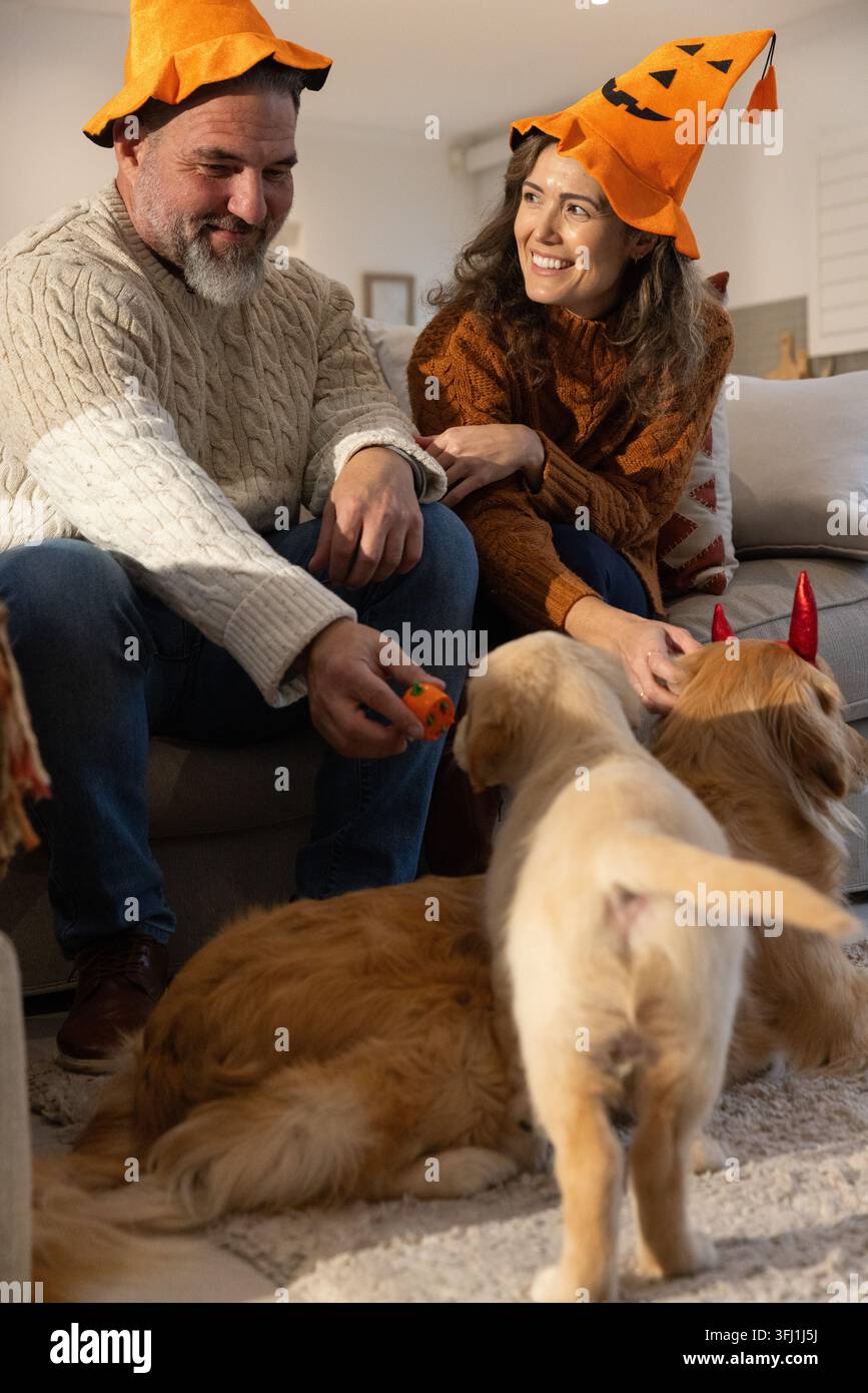 Couple assis sur un canapé portant des chapeaux en citrouille, tenant un jouet citrouille, caressant un chien avec des cornes rouges. Confortable, lanterne d’Halloween, citrouille, automne Banque D'Images