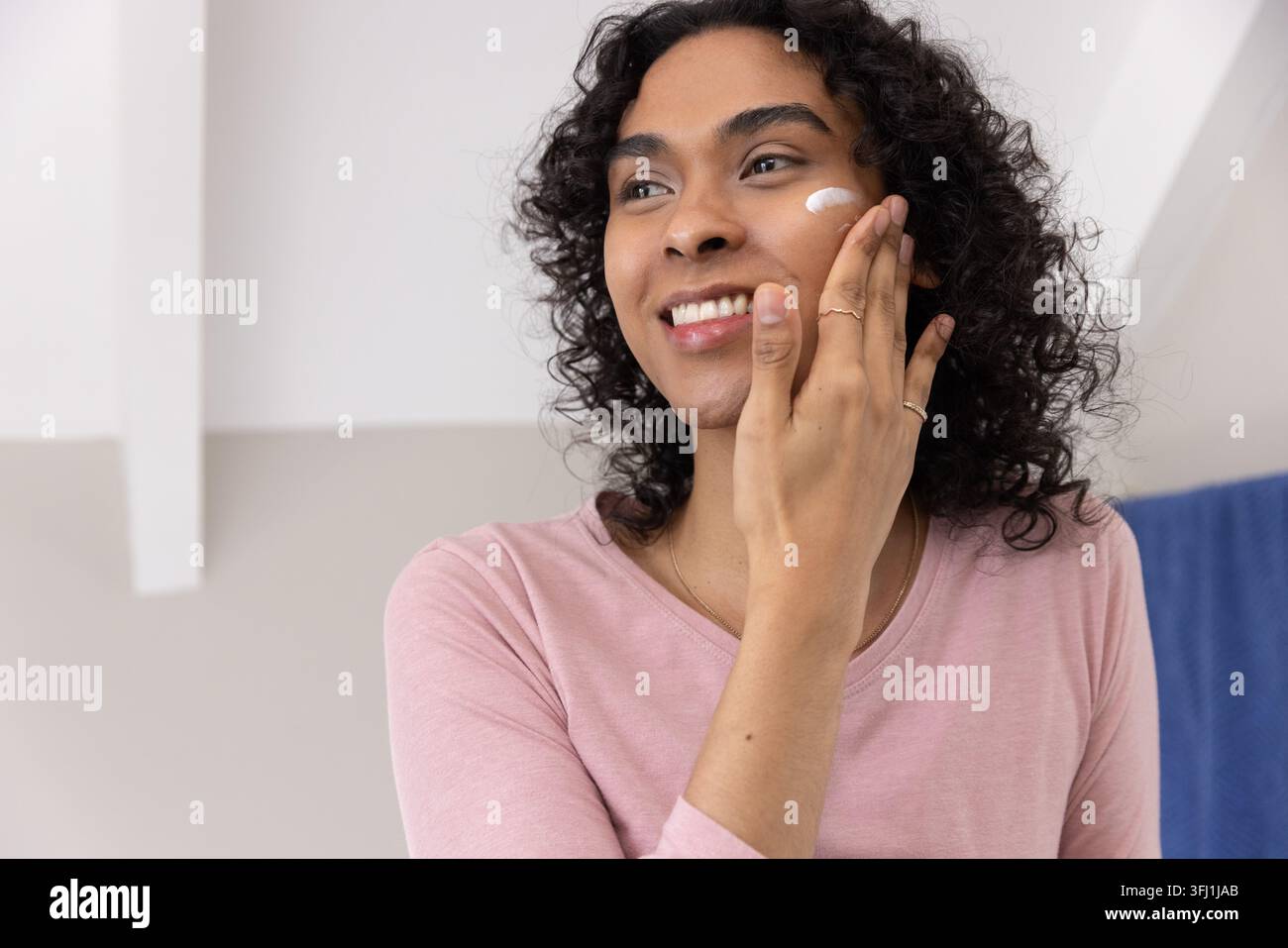 Personne non binaire appliquant la crème blanche à la joue à la vanité, portant le dessus rose, les bagues et le collier. Hydratant, soin de la peau, soin de soi, cheveux frisés, lumière douce Banque D'Images