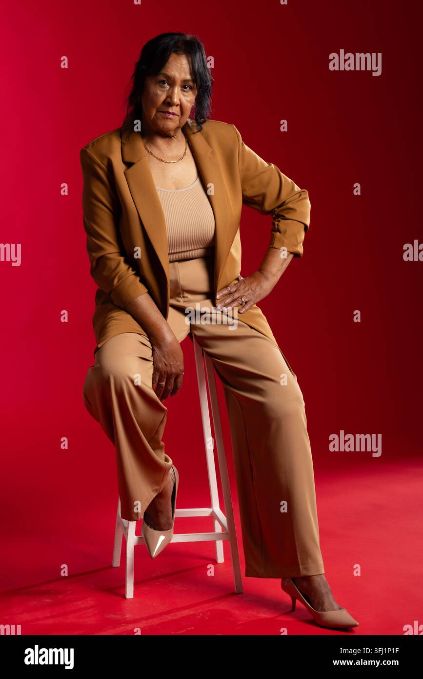 Sénior afro-américaine assise sur un tabouret en bois blanc posant en studio portant un blazer beige Banque D'Images