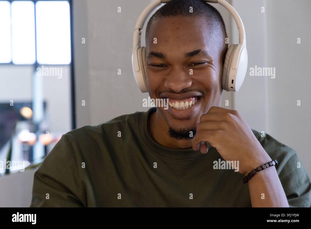 Homme adulte afro-américain souriant au bureau dans le bureau avec casque blanc et bracelet par menton. Portrait, casque, barbu, olive, vert, coworking, Banque D'Images