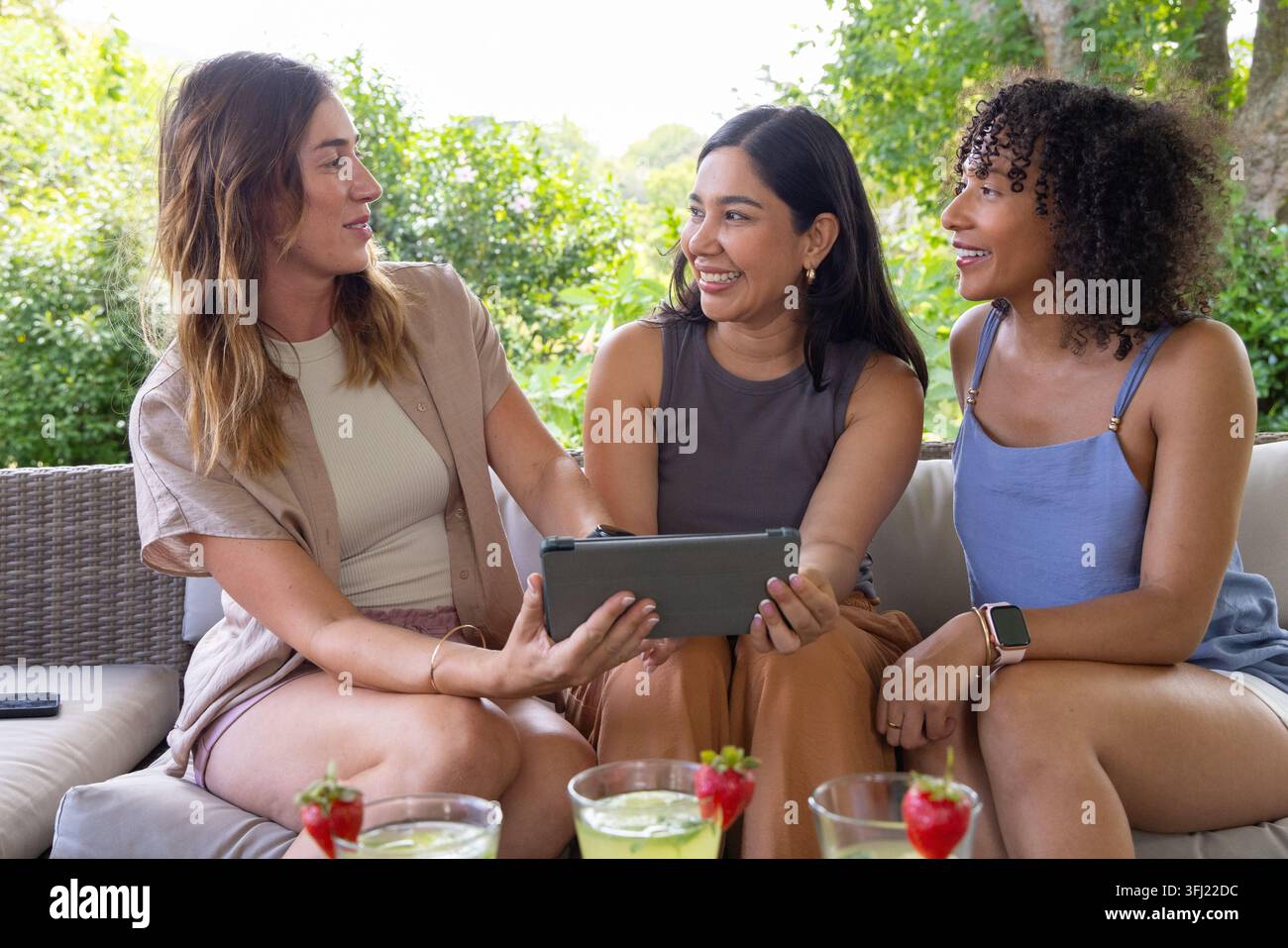 Trois amies diverses assises côte à côte sur un canapé de terrasse, regardant une tablette et sirotant des boissons vertes. Extérieur, arrière-cour, femmes, c Banque D'Images