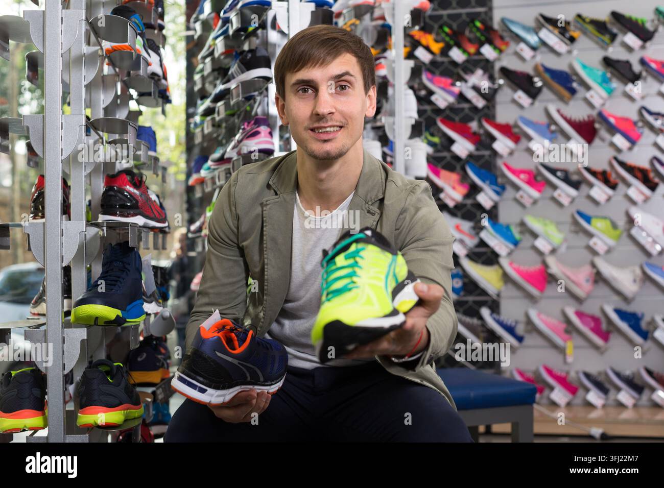 Jeune homme joyeux tenant une chaussure de course vibrante avec un sourire confiant dans un magasin de sport, confirmant son choix final parmi de nombreux modèles Banque D'Images
