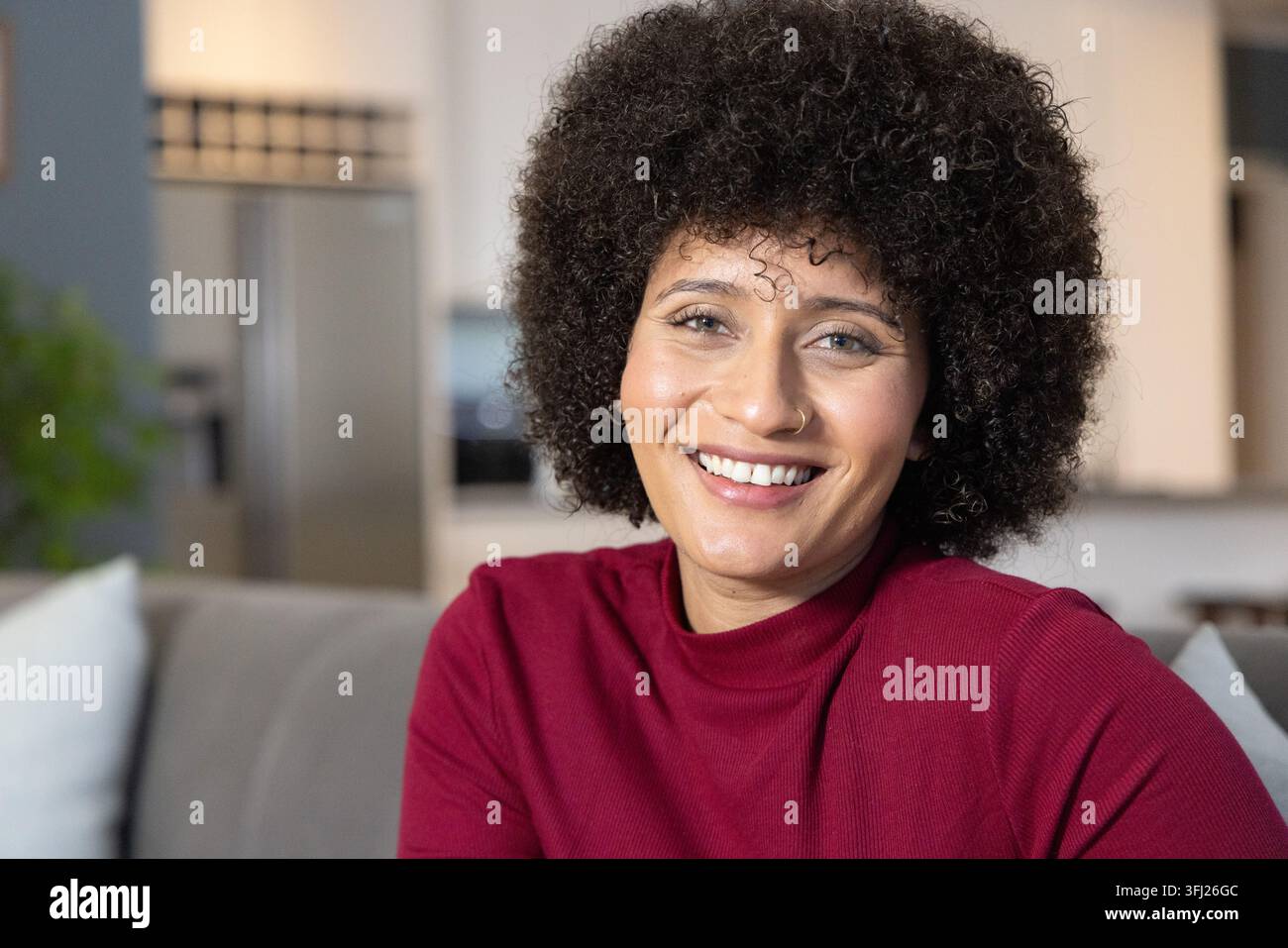 Femme afro-américaine assise sur un canapé gris avec un réfrigérateur flou en arrière-plan, portant un haut marron, souriante. Portrait, cosy, moderne, ope Banque D'Images