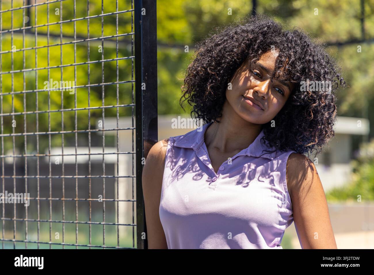 Femme afro-américaine appuyée contre un poteau métallique près d'une clôture, la lumière du soleil projetant des ombres sur un polo lilas. Extérieur, parc, ensoleillé, décontracté Banque D'Images