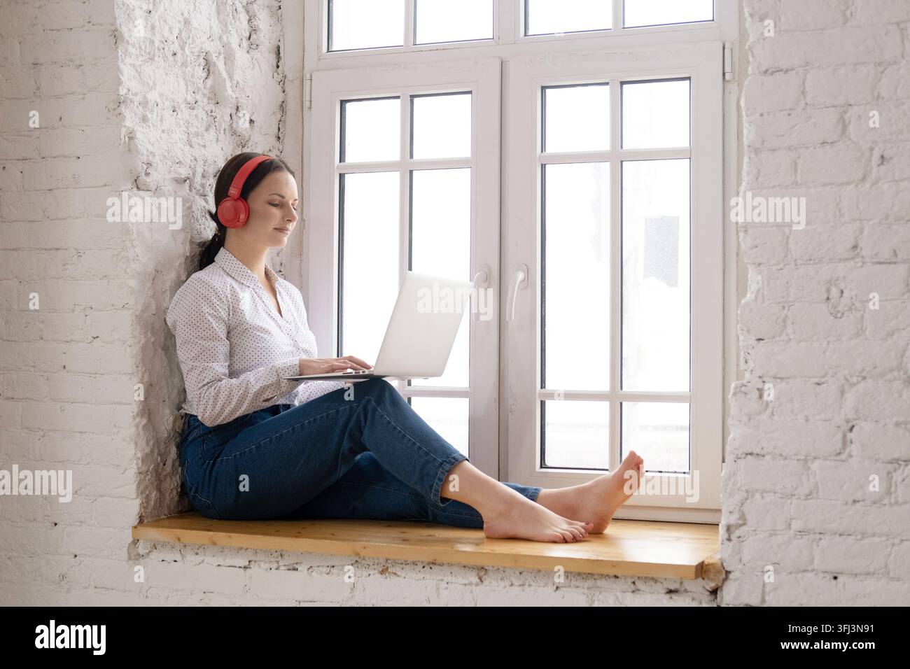 Jeune femme assise à la fenêtre à la maison avec un ordinateur portable et un casque Banque D'Images