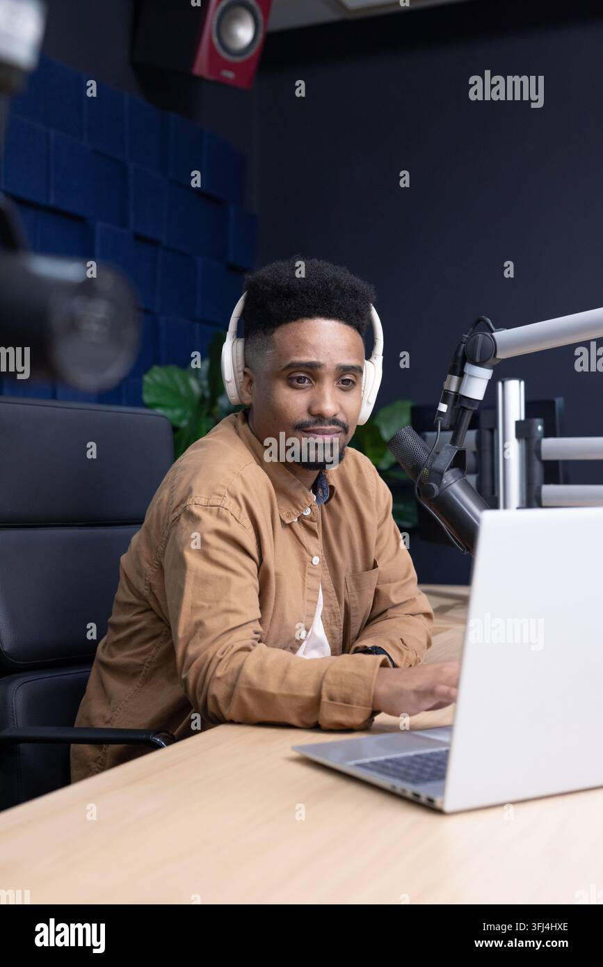 Homme afro-américain en train d'enregistrer à un bureau en bois dans un studio, portant des casques, utilisant un ordinateur portable et un micro Banque D'Images