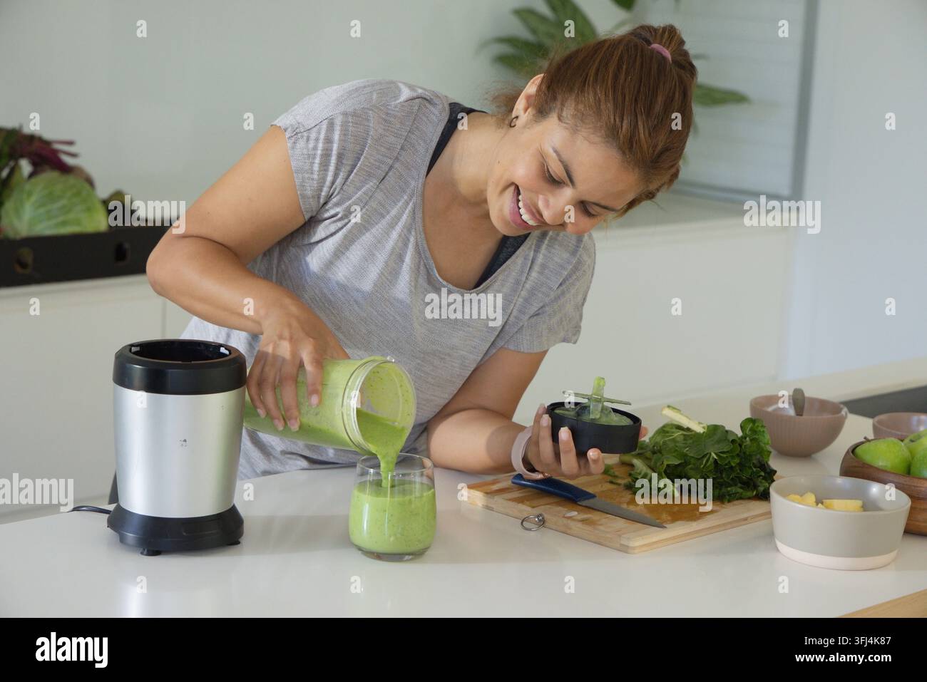 Femme versant un smoothie vert vif dans un verre court sur un comptoir de cuisine blanc, tenant le couvercle du mixeur. Femelle, chef à domicile, éclairé par le soleil, mo Banque D'Images