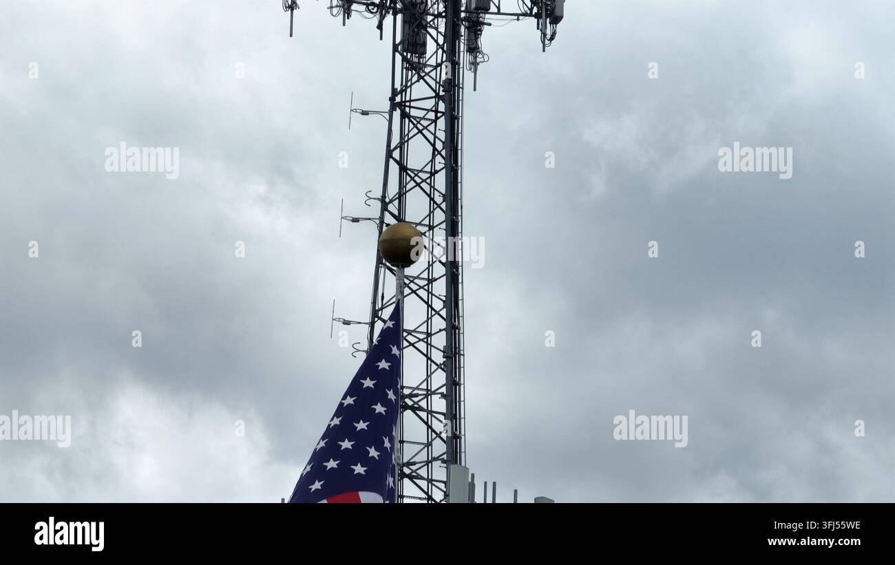 Un drapeau américain fier flotte au vent devant une antenne relais s'élevant dans un ciel nuageux. Symbole puissant de liberté, de communication moderne et de technologie Banque D'Images