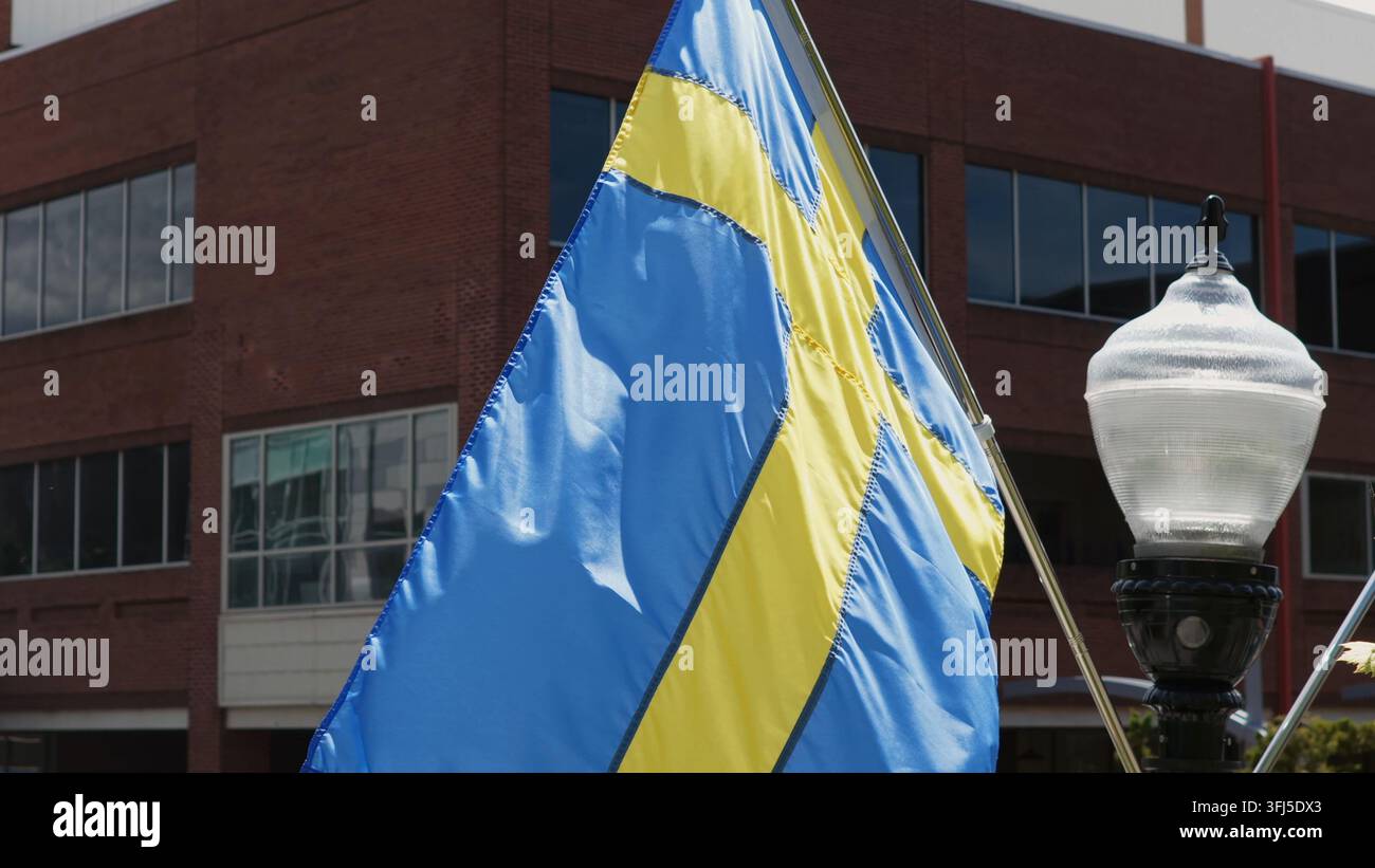 Le drapeau suédois flotte à côté d'un lampadaire vintage devant un bâtiment historique en briques dans une petite ville américaine, États-Unis, marquant les célébrations de la Coupe du Monde 2026 au milieu de la forêt Banque D'Images