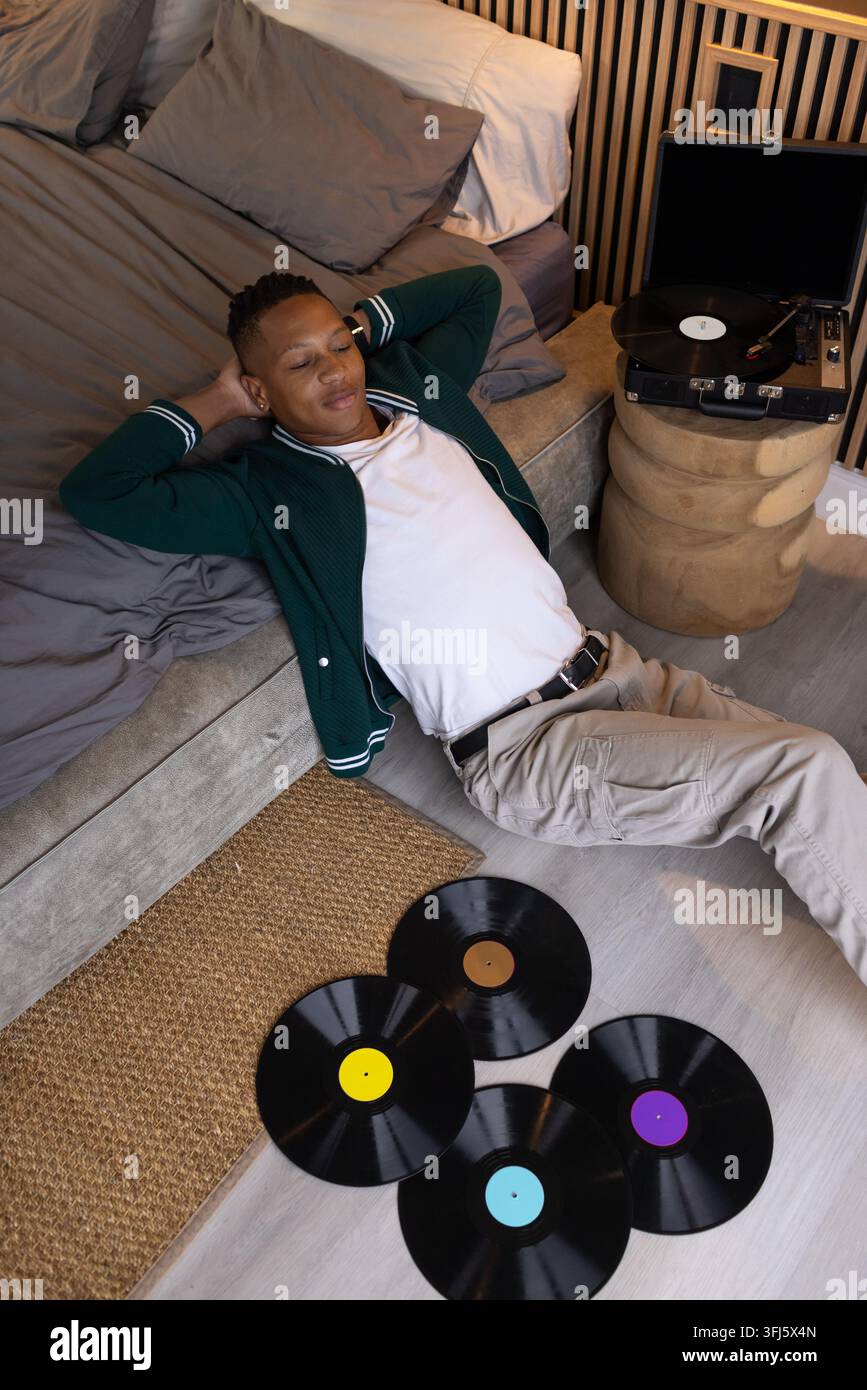 Homme africain dans la vingtaine allongé sur le sol de la chambre à côté d'un lit bas, platine vinyle en lecture Banque D'Images