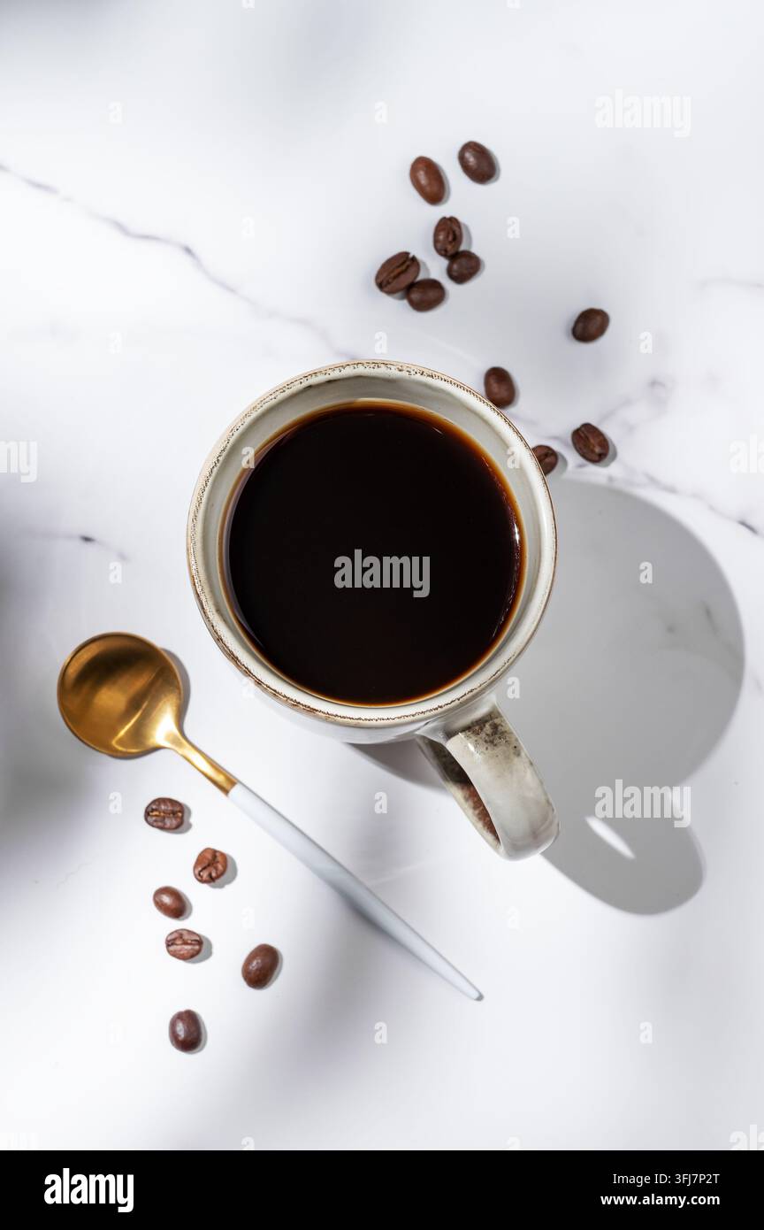 Café espresso fraîchement préparé dans une tasse sur un fond de marbre blanc avec des grains de café éparpillés, une cuillère et la lumière du matin. Un petit-déjeuner délicieux co Banque D'Images