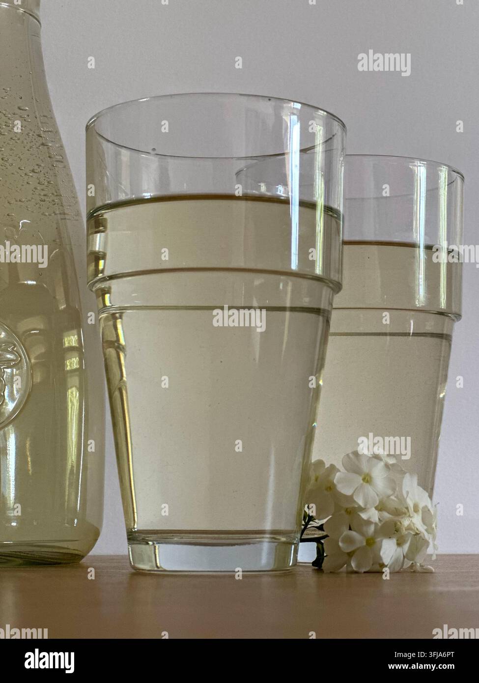 Deux verres de boisson claire avec une bouteille et des fleurs blanches sur une table en bois. Deux grands verres clairs remplis d'une boisson légère à côté d'un verre b - Image de stock capturée avec un smartphone