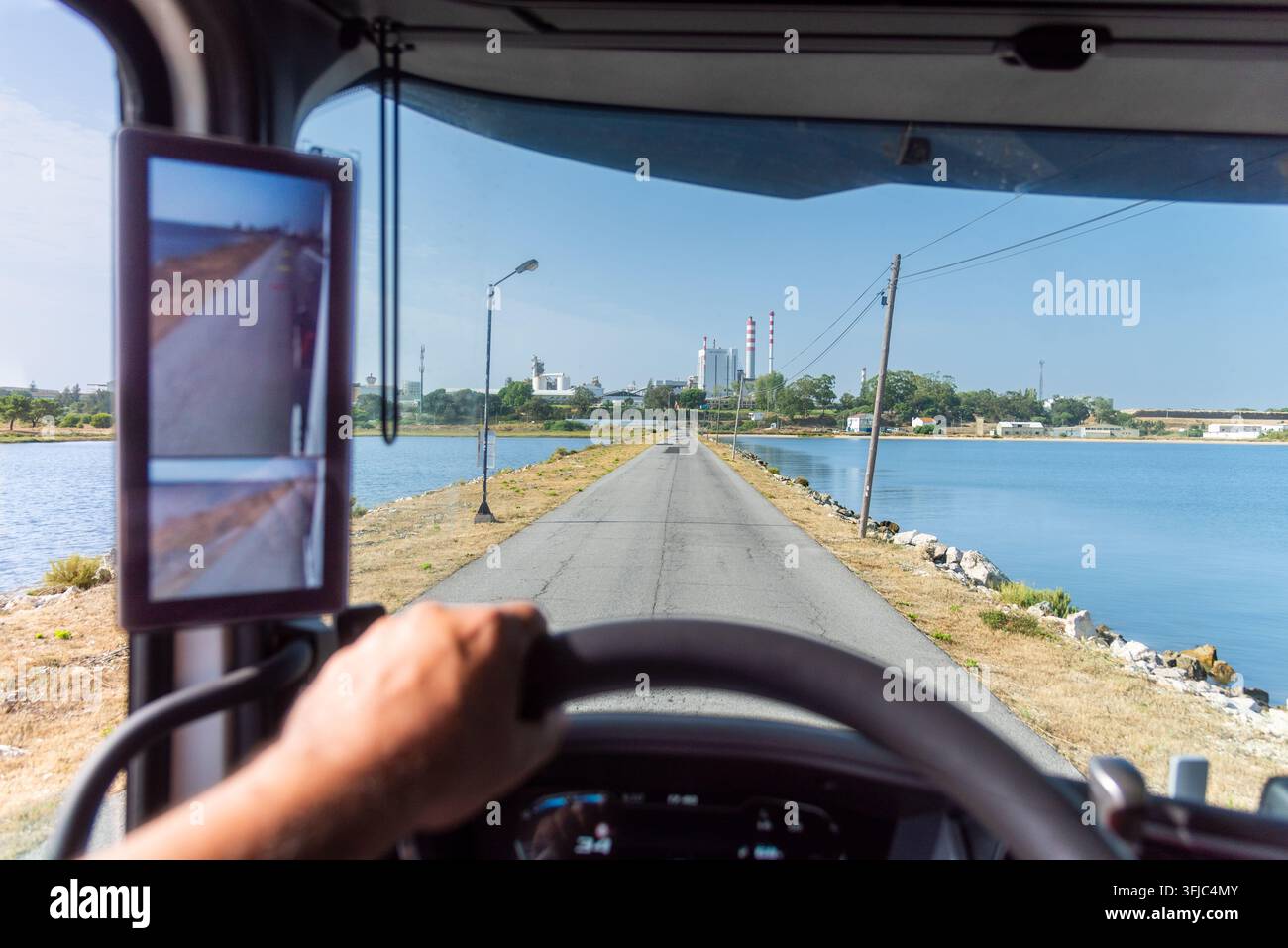 Vue depuis la cabine d'un camion circulant sur une route construite sur une chaussée entourée d'eau, avec une installation industrielle en arrière-plan. Banque D'Images