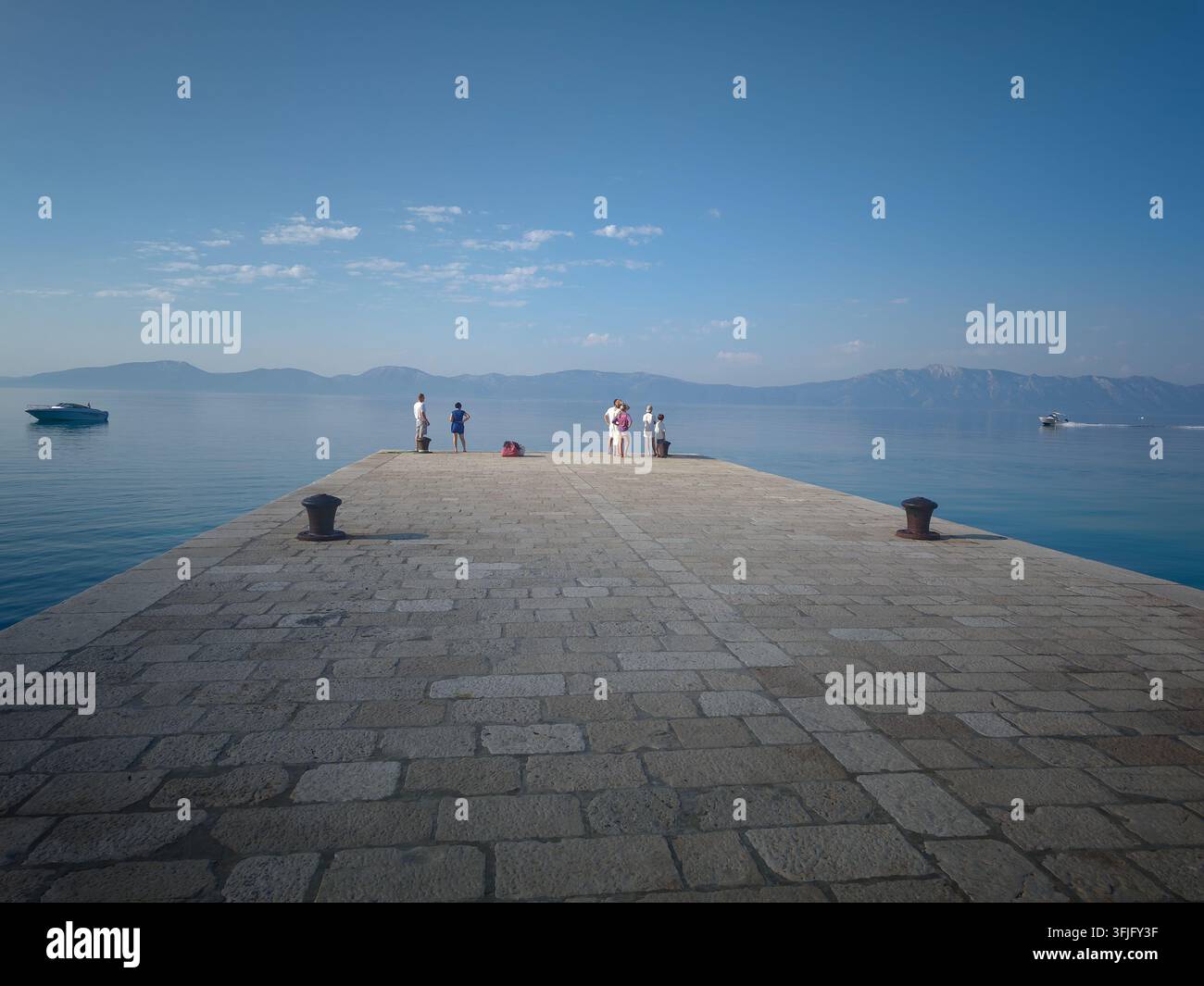 Un groupe de touristes se tient au bout d'une jetée en pierre, admirant la mer bleue calme et les montagnes lointaines. - Image de stock capturée avec un smartphone