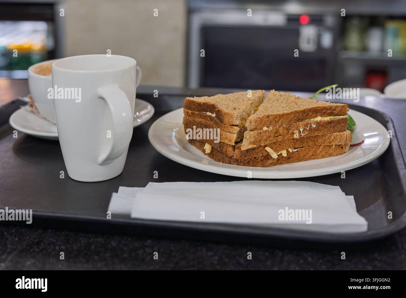 Deux moitiés triangulaires d'un sandwich au pain complet avec garniture au fromage visible, servi sur une assiette blanche accompagné d'une grande tasse de café blanche, de serviettes blanches fo Banque D'Images