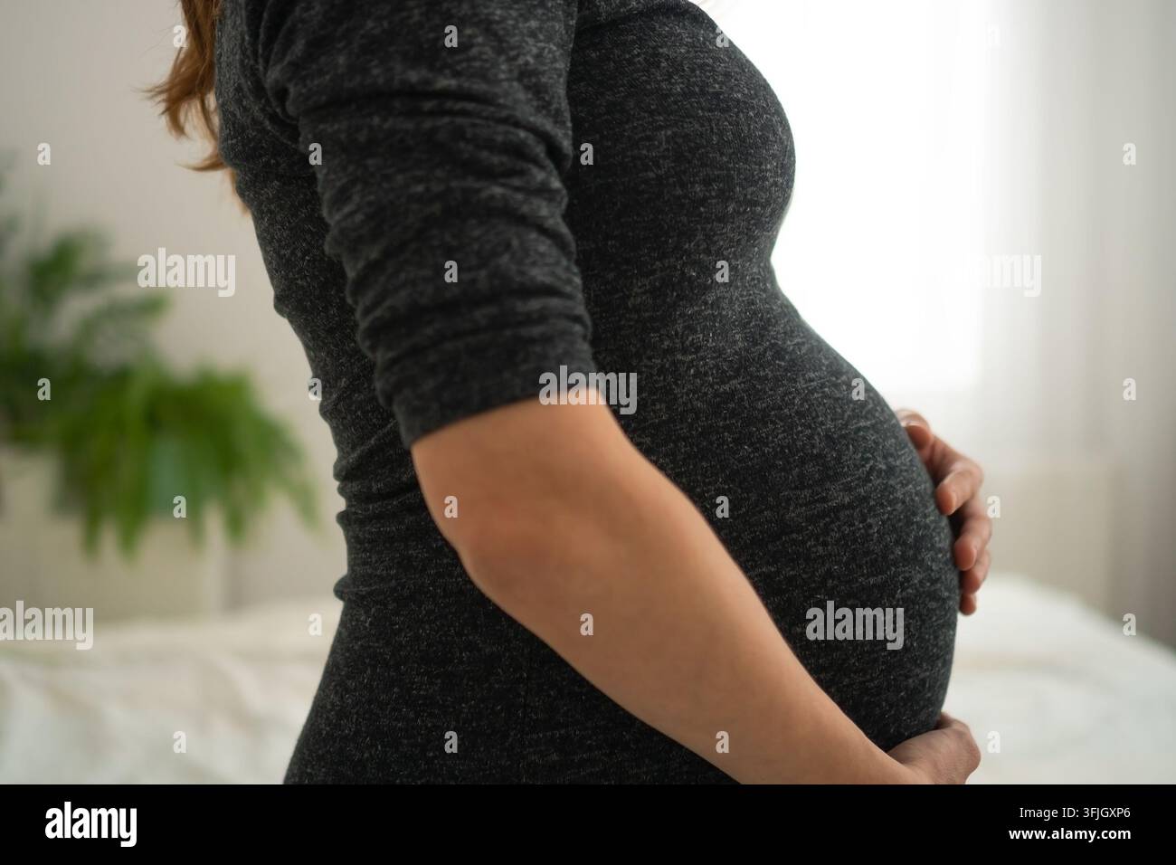Close-up de profil d'une femme enceinte tenant doucement son ventre rond dans une chambre sereine. Mère enceinte berçant son ventre en vue latérale tendre. Future maman touchant son ventre avec un soin intime à côté d'un lit blanc Banque D'Images