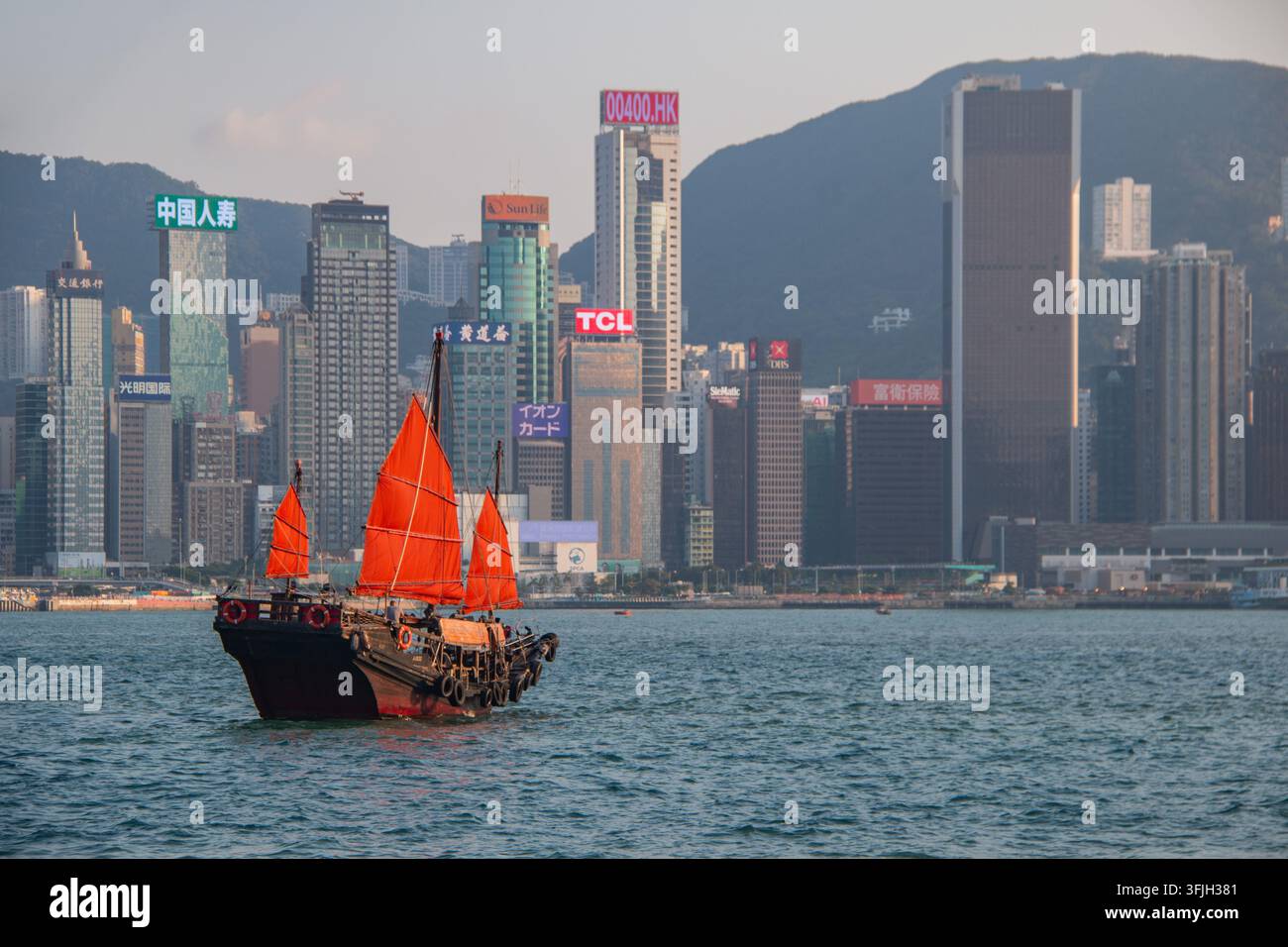 Bateau junk chinois traditionnel avec des voiles rouges vives traversant le Victoria Harbour devant l'horizon emblématique de l'île de Hong Kong au coucher du soleil Banque D'Images