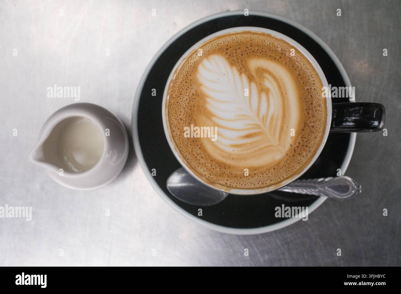 Un barista crée un art latte en forme de feuille complexe dans une tasse noire accompagnée d'une crème sur une table en métal dans un café. Banque D'Images
