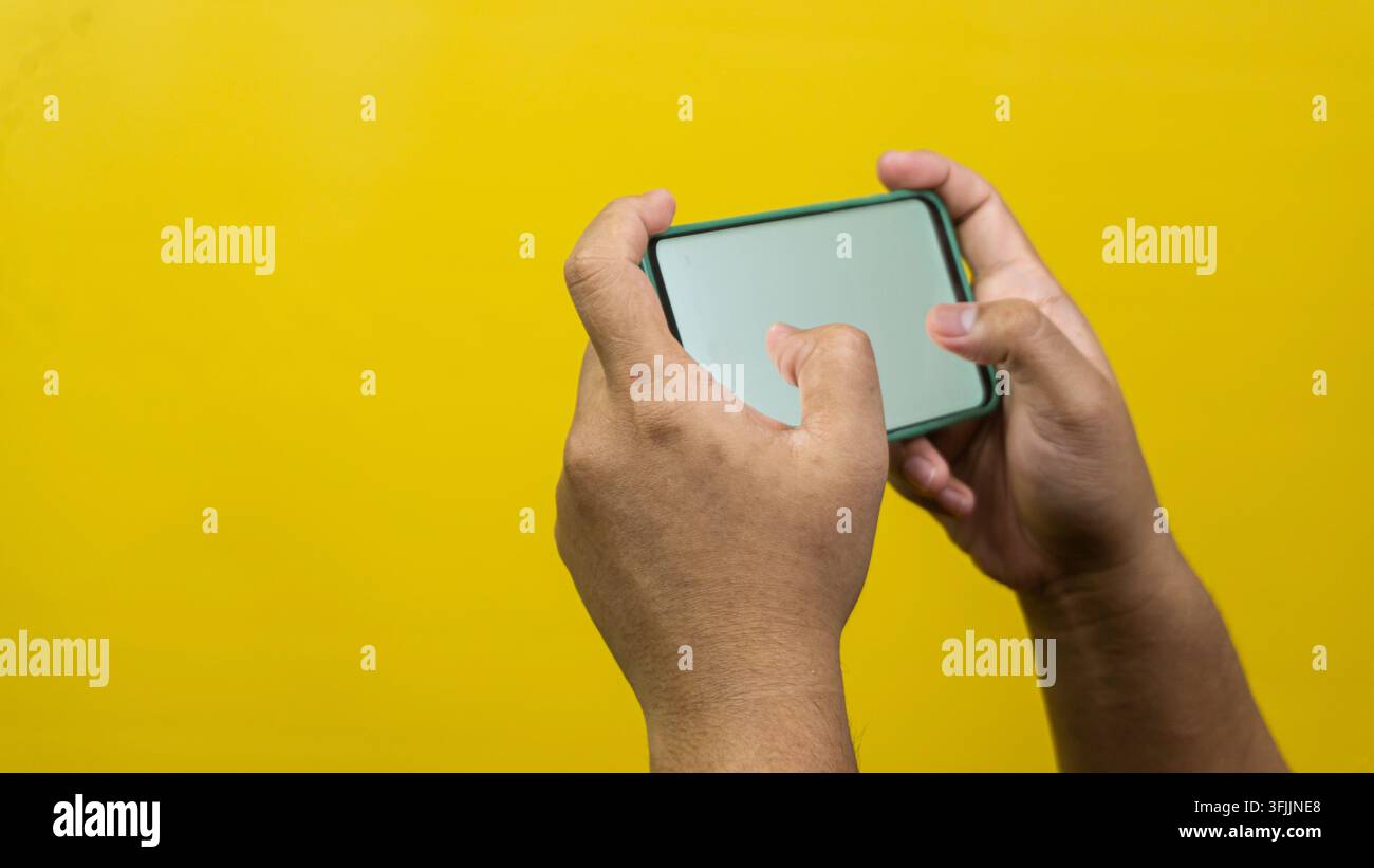 Deux mains d'hommes adultes saisissent un smartphone vierge horizontalement sur un fond jaune uni, démontrant des jeux mobiles actifs ou une saisie de texte rapide p Banque D'Images