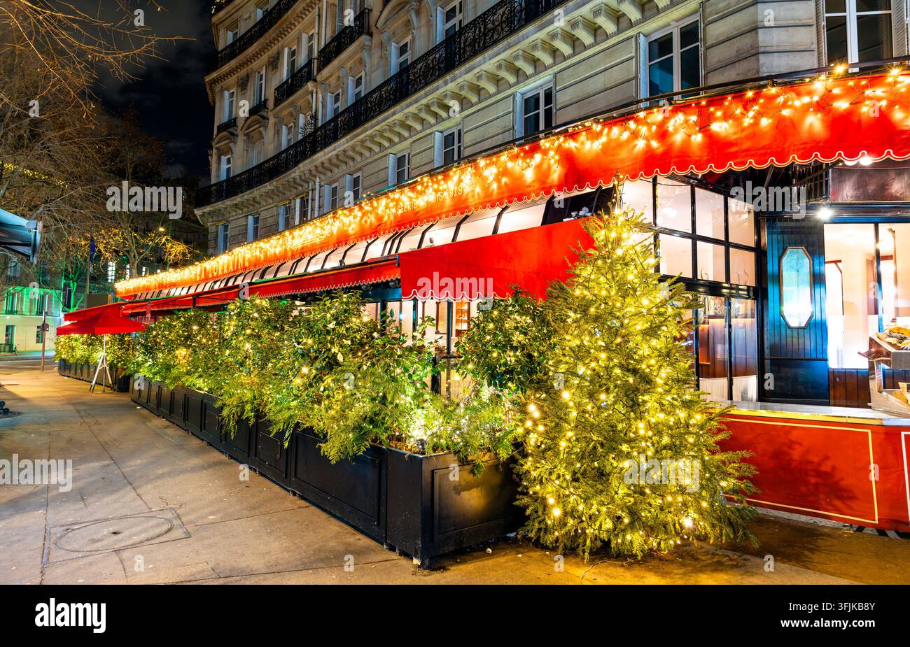 Une terrasse de restaurant parisienne typique décorée d'arbres de Noël illuminés et de guirlandes lumineuses le long d'un trottoir la nuit à Paris, France. Banque D'Images