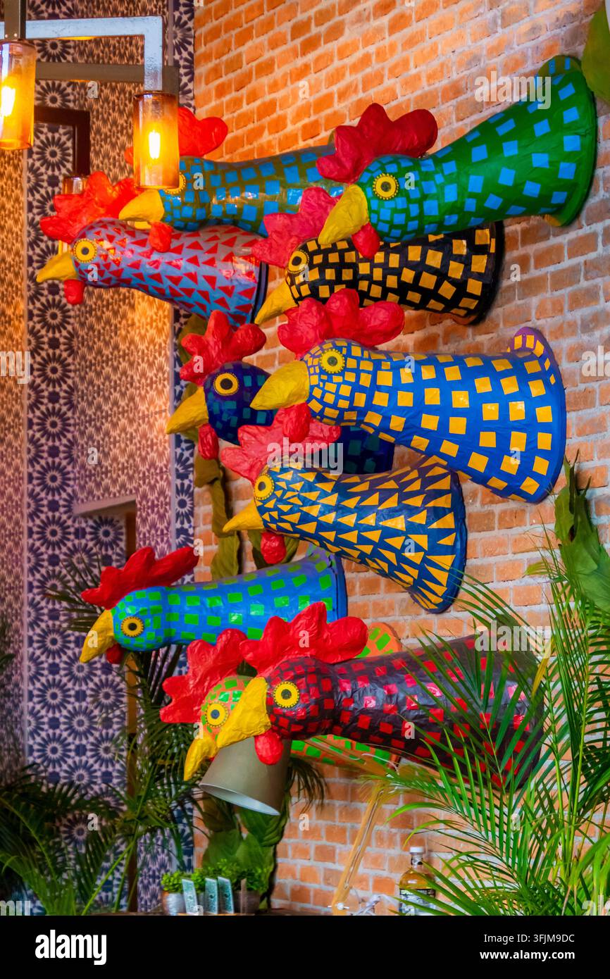 Décorations murales colorées en forme de coq sur un mur de briques apparentes avec des lumières suspendues chaudes et des plantes tropicales vertes dans un intérieur de restaurant animé Banque D'Images