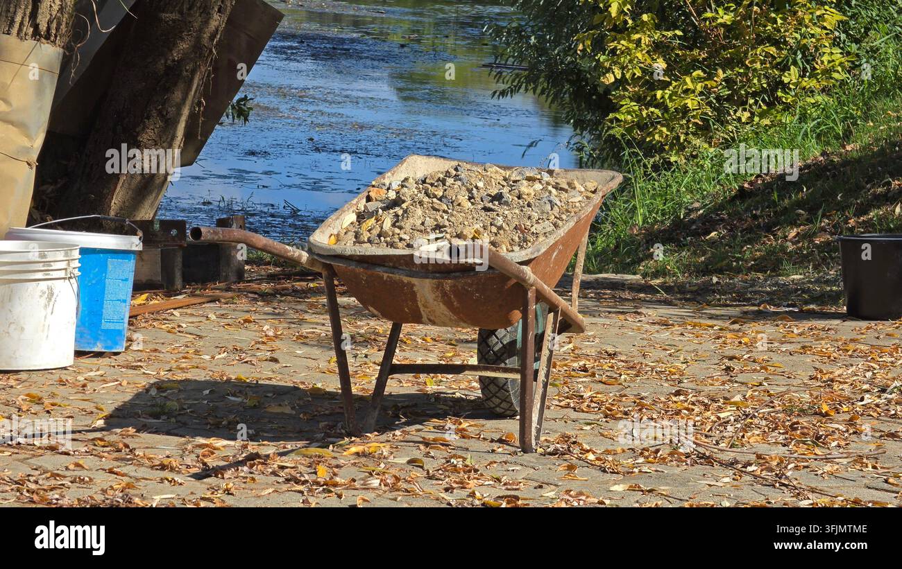 Une vue d'une brouette métallique vieillotte chargée de pierres de gravier lourdes et de terre garée sur un trottoir pavé de briques recouvert de feuilles d'automne tombées à côté Banque D'Images