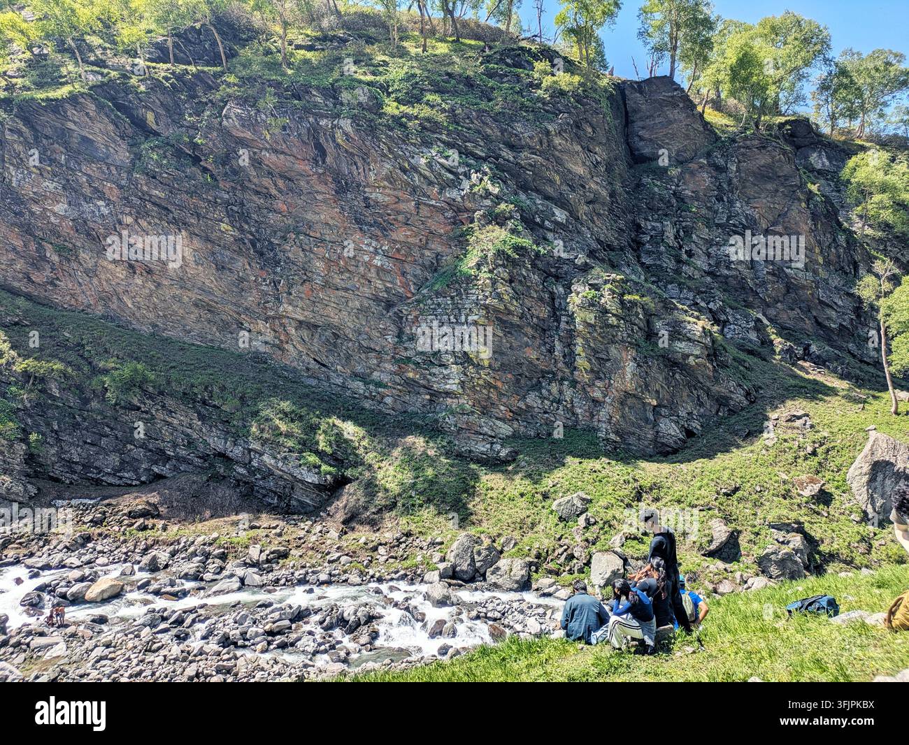 Trekkeurs se reposant près d’un ruisseau de montagne qui coule sous une falaise rocheuse massive sur la route du col de Hampta, Inde - Image de stock capturée avec un smartphone