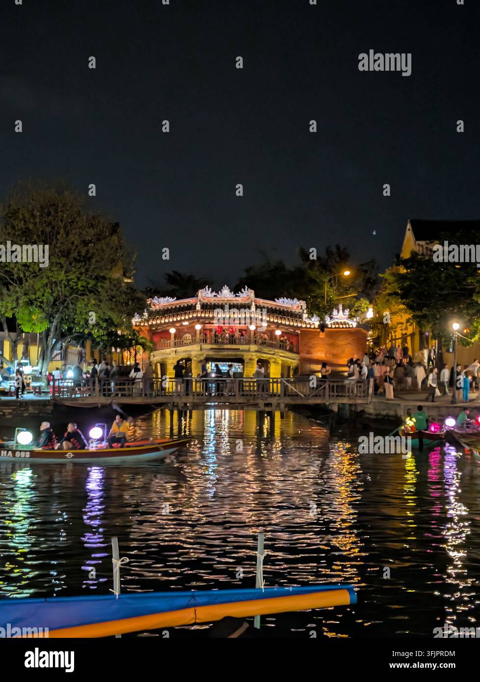 HOI AN, VIÊT NAM - 10 MARS 2026 : Vue nocturne du pont couvert japonais historique illuminé au-dessus de la rivière avec des bateaux de touristes - Image de stock capturée avec un smartphone