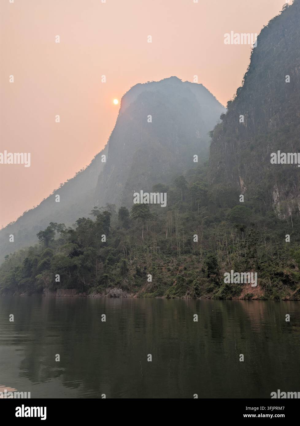 Vue panoramique du disque solaire lumineux disparaissant derrière un immense pic karstique couvert de jungle enveloppé de brume, avec des eaux fluviales paisibles au premier plan. - Image de stock capturée avec un smartphone