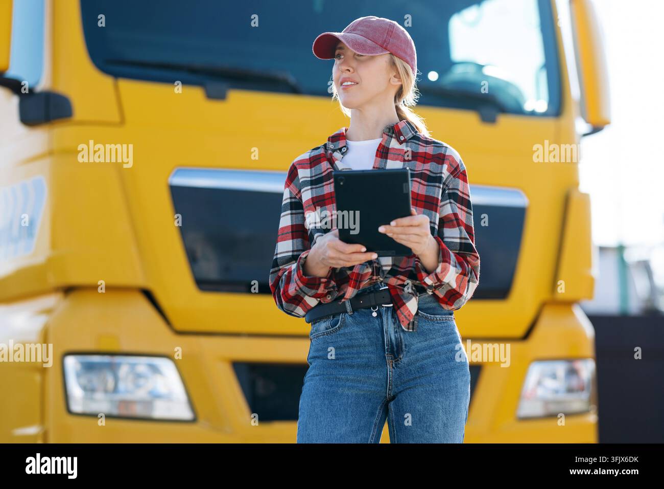 Une femme portant une chemise à carreaux et une casquette de baseball tient une tablette numérique tout en travaillant à l'extérieur près d'un véhicule de transport lourd pendant la journée Banque D'Images