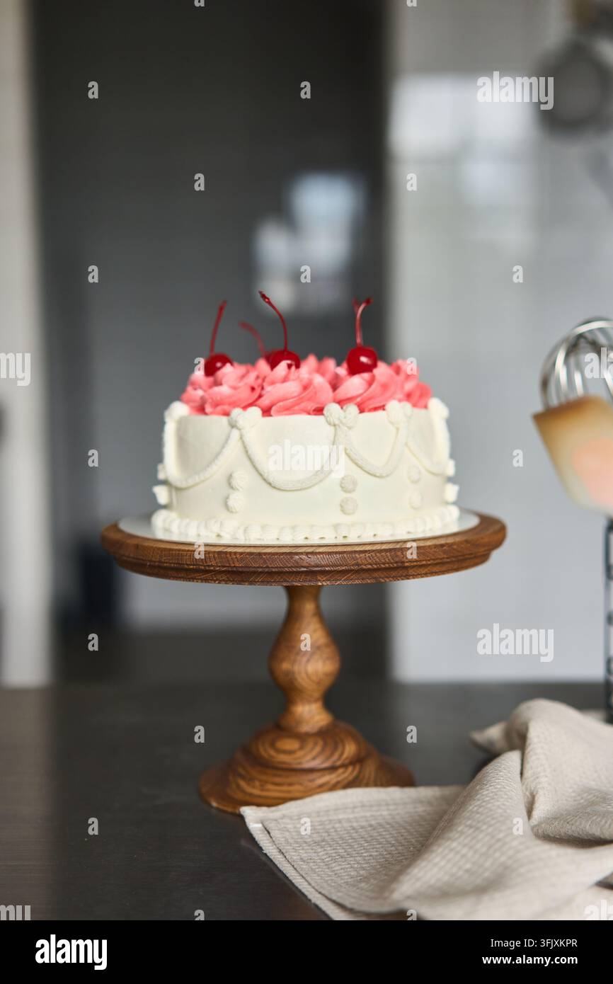 Gâteau de fête rond et blanc avec un glaçage en rosettes de crème au beurre rose et une garniture de cerises sur un piédestal en bois sculpté, photographié dans une cuisine lumineuse avec lumière naturelle Banque D'Images