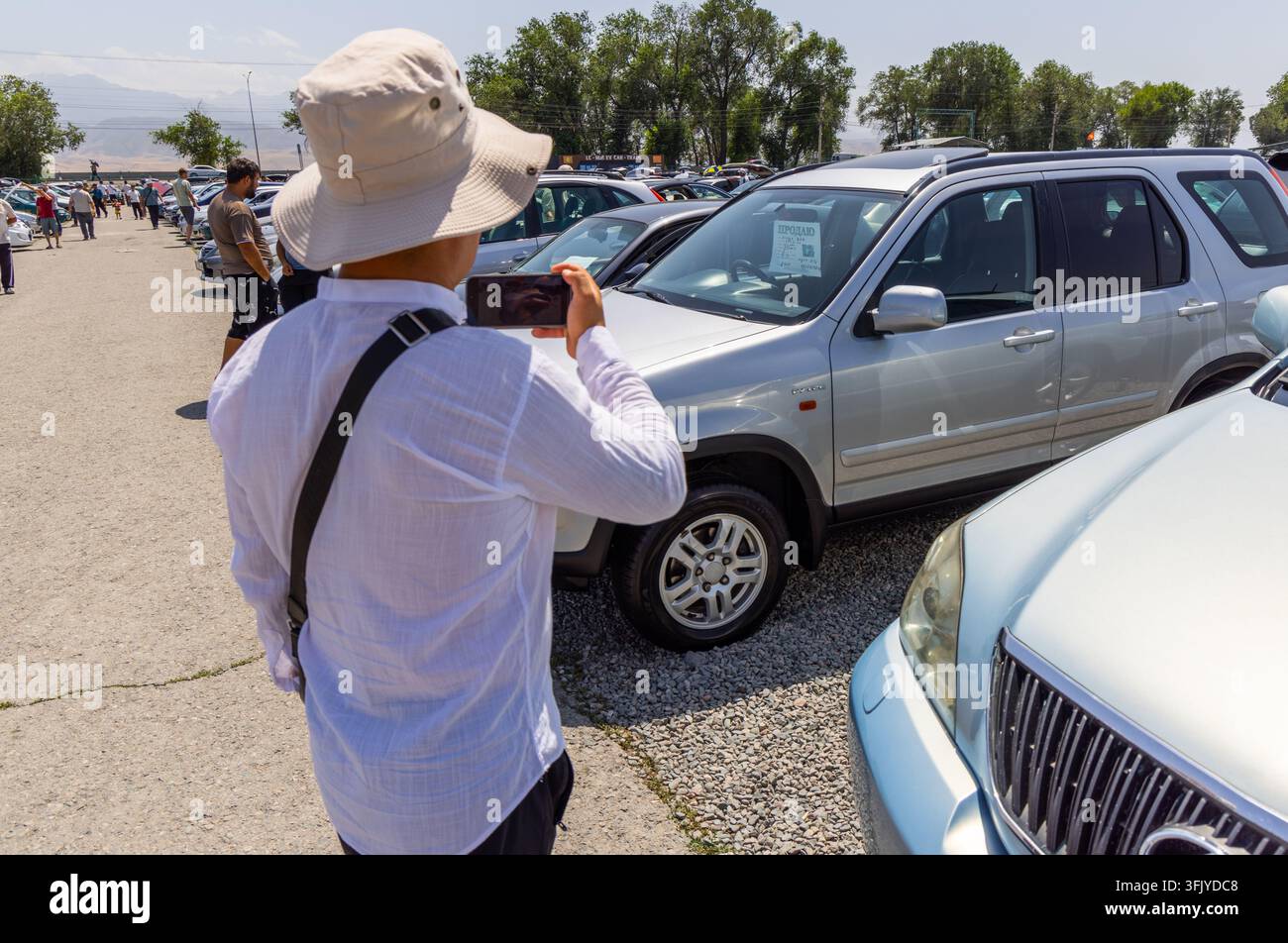 Bichkek, Kirghizistan - 29 juin 2025 : Une personne portant un chapeau à larges bords photographie un SUV argenté sur un marché de voitures d'occasion en plein air. Des rangées de véhicules sont garées Banque D'Images
