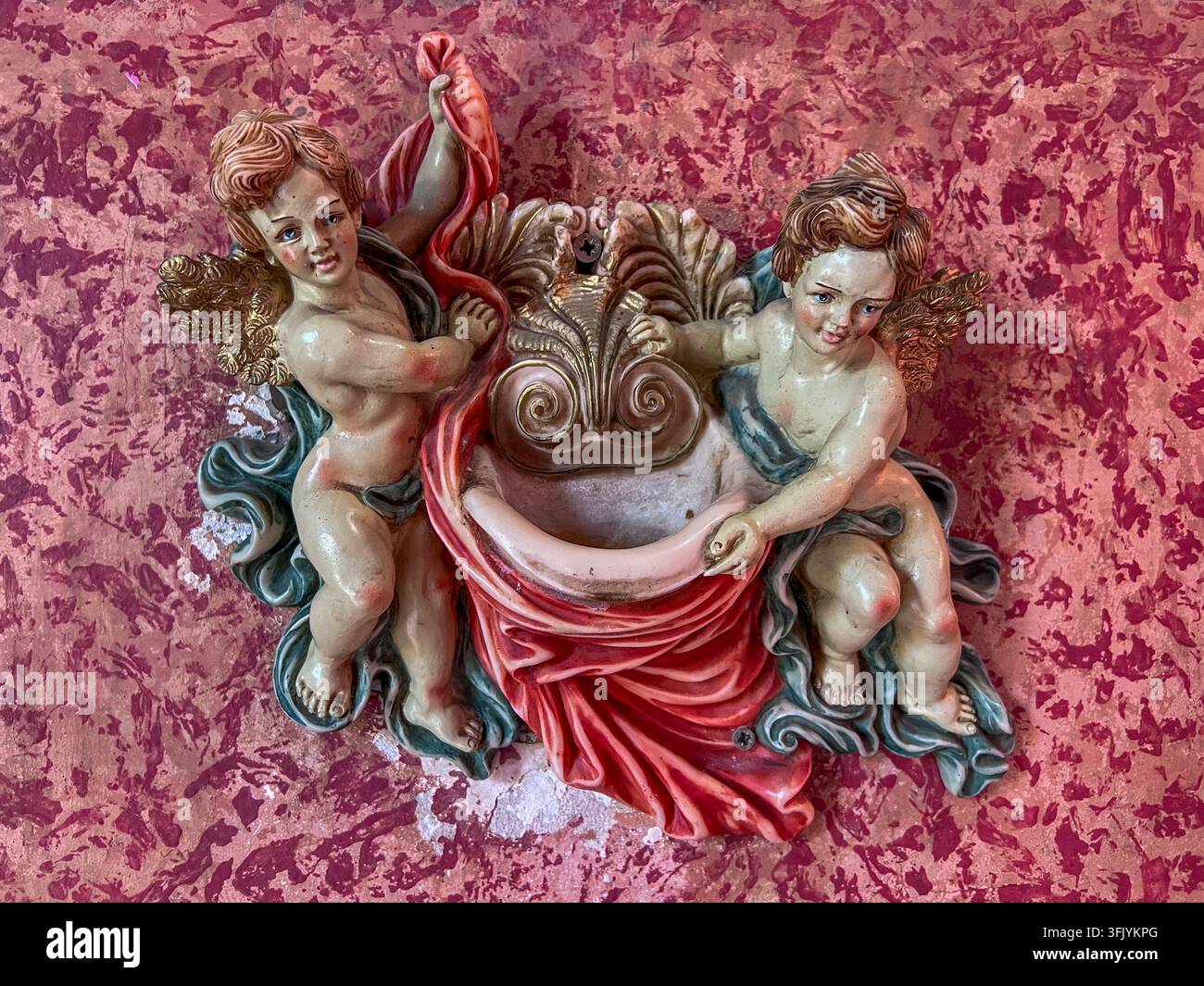 Fontaine d'eau bénite baroque ornée de deux figures de chérubins peintes et d'une coquille dorée, fixée sur un mur en marbre rouge à l'intérieur d'une église - Image de stock capturée avec un smartphone