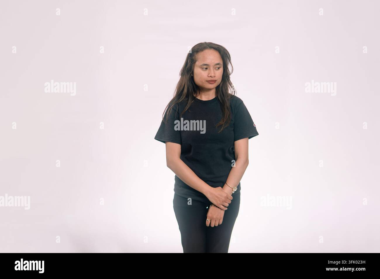 Portrait d'une femme asiatique timide en t-shirt noir, debout les mains jointes et regardant sur le côté sur un fond blanc. Banque D'Images