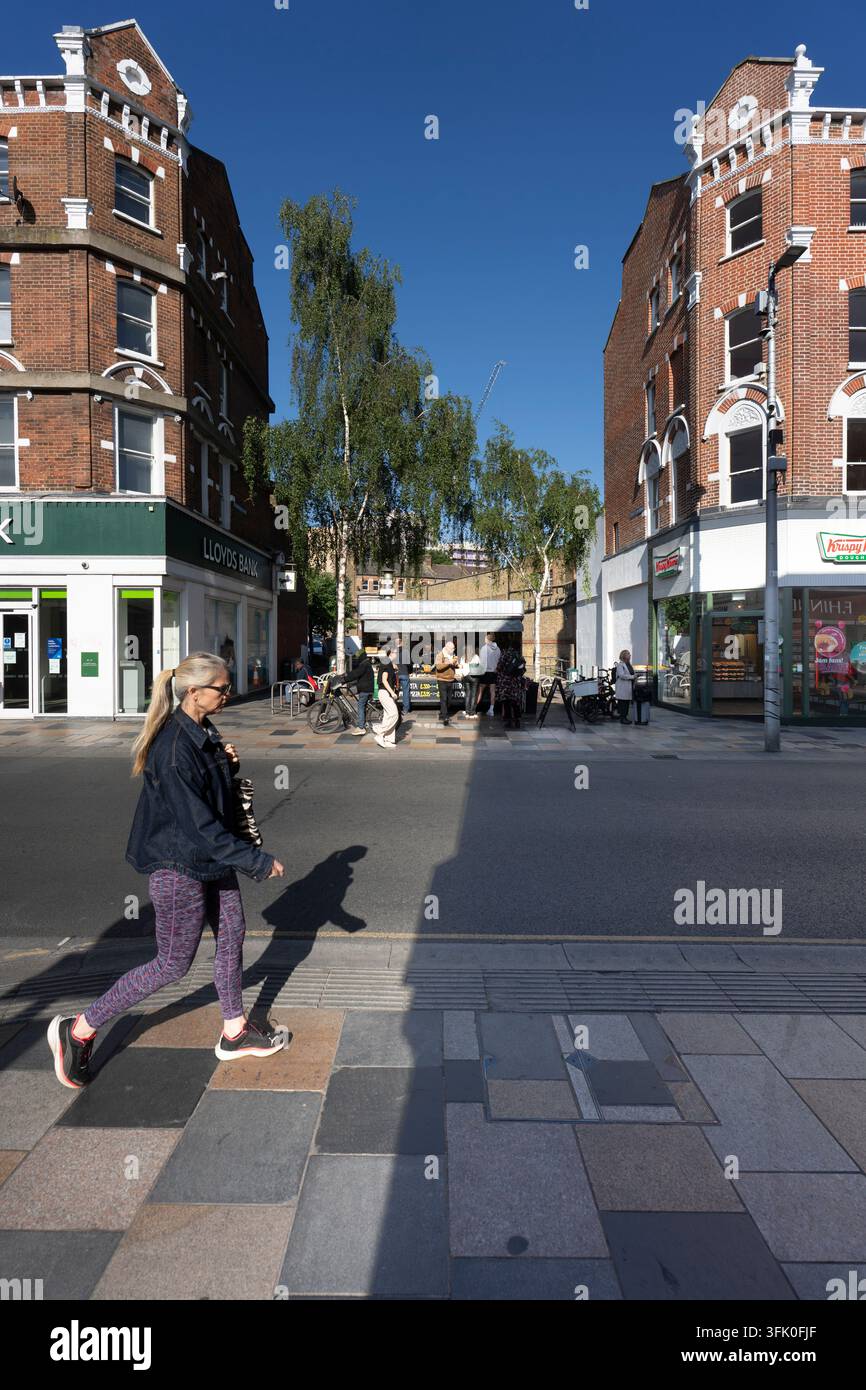 Une femme passe devant des boutiques sur St. John's Hill près de Clapham Junction, à Londres, tandis que d'autres se rassemblent à un stand de nourriture par une journée ensoleillée. Banque D'Images