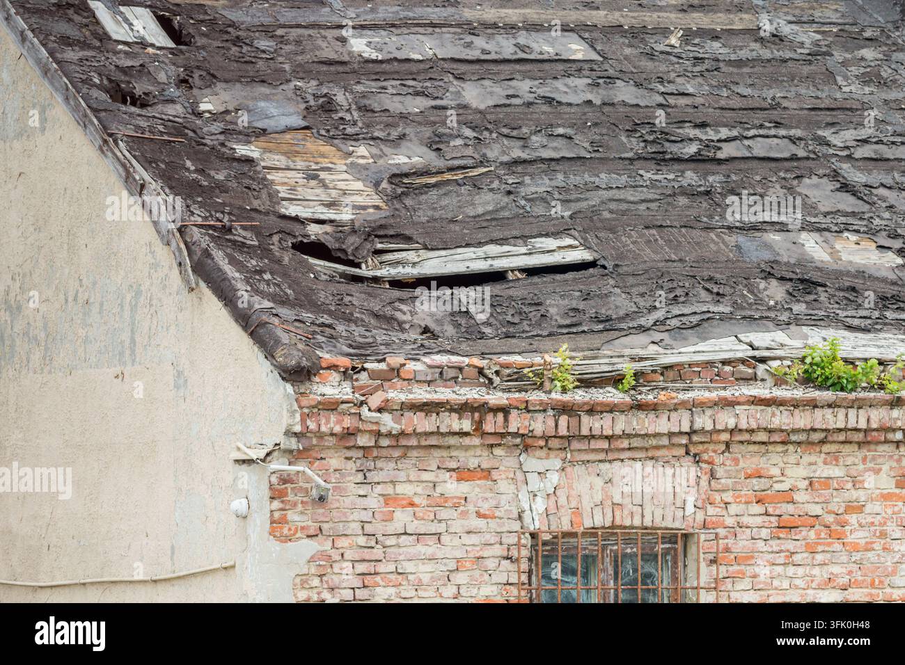 Ancien toit érodé avec des matériaux de toiture endommagés au-dessus d'un mur de briques rouges délabré où la végétation pousse le long du sommet. Banque D'Images