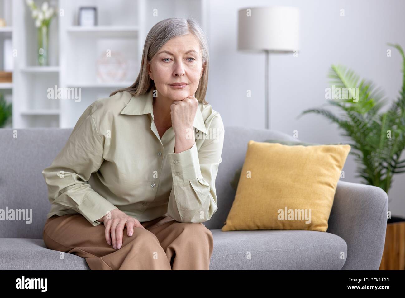 Femme âgée assise sur un canapé, regardant pensivement et au loin, faisant face à la solitude, au chagrin et à l'anxiété à la maison, réfléchissant à la perte et à la lutte émotionnelle dans une solitude tranquille Banque D'Images