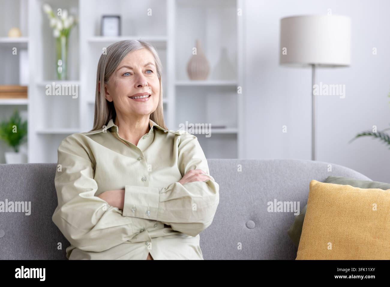 Femme mûre aux cheveux gris assise sur un canapé lumineux, souriant et regardant au loin, profitant d'un moment paisible et satisfait de contemplation détendue et de bien-être confiant à la maison Banque D'Images
