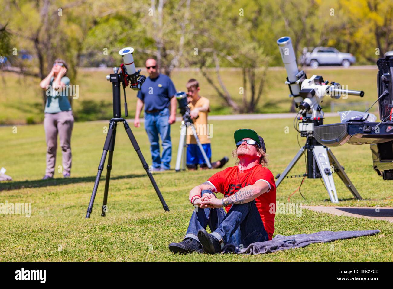 Arkansas, États-Unis, 08 avr. 2024 – Un homme en chemise rouge est assis sur l'herbe tandis que d'autres utilisent des télescopes pour des observations astronomiques par une journée ensoleillée Banque D'Images