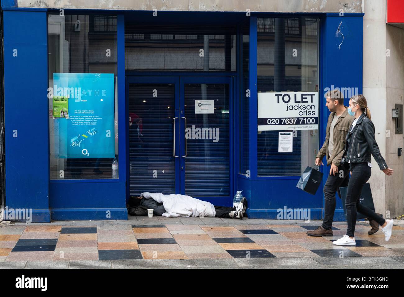 Une personne sans-abri dort dans l'entrée d'une boutique vacante sur St John's Road, Battersea, Londres, en raison de la pauvreté, tandis que des acheteurs passent à côté. Banque D'Images