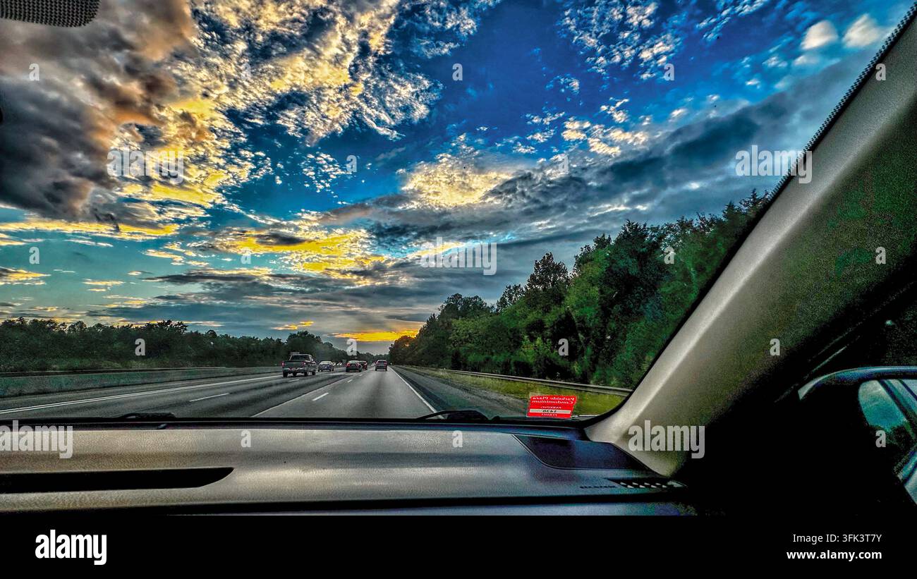 La vue depuis l'intérieur d'une voiture roulant sur une autoroute à plusieurs voies lors d'un coucher de soleil dramatique avec des nuages texturés et colorés. Banque D'Images