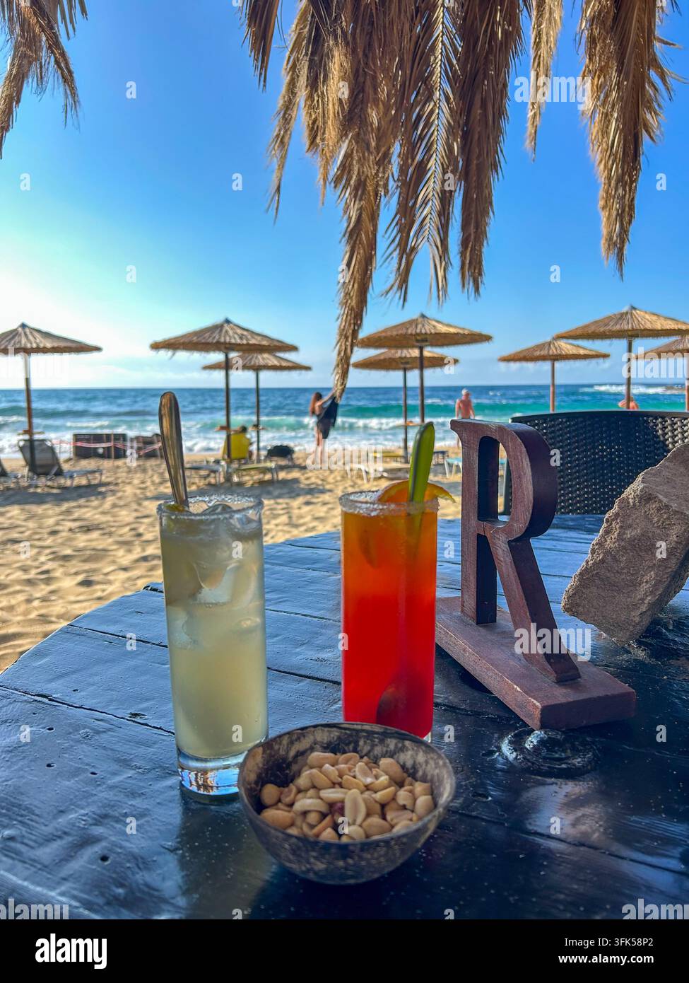 Deux cocktails rafraîchissants et des cacahuètes sur une table de bar de plage, avec des parasols en paille baignés de soleil et la mer en arrière-plan, Stavros, Crète. - Image de stock capturée avec un smartphone
