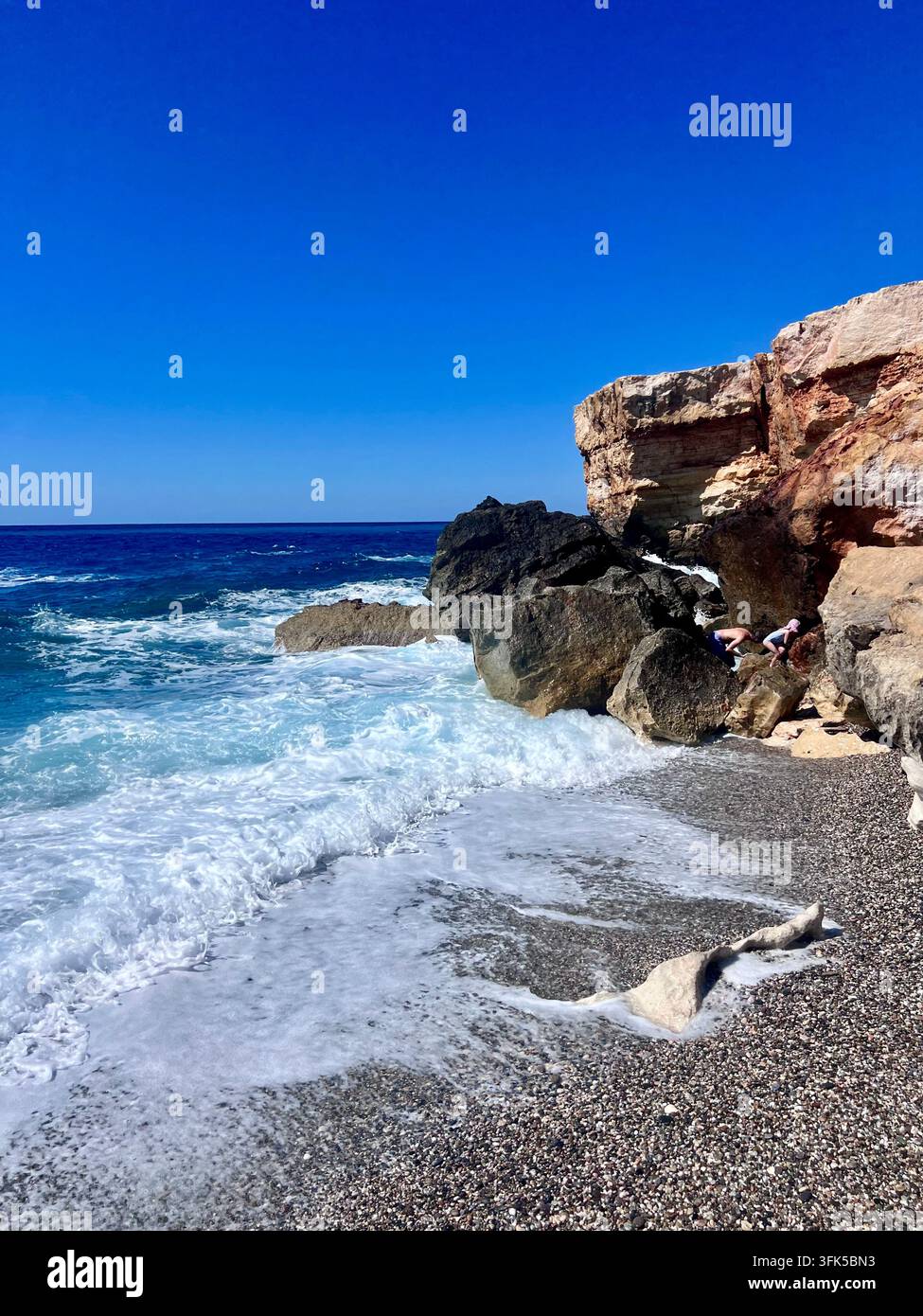 Deux personnes explorent les criques rocheuses sous des falaises superposées à la plage de Spilies, avec des vagues écumeuses déferlant sur des galets, Crète, Grèce. - Image de stock capturée avec un smartphone