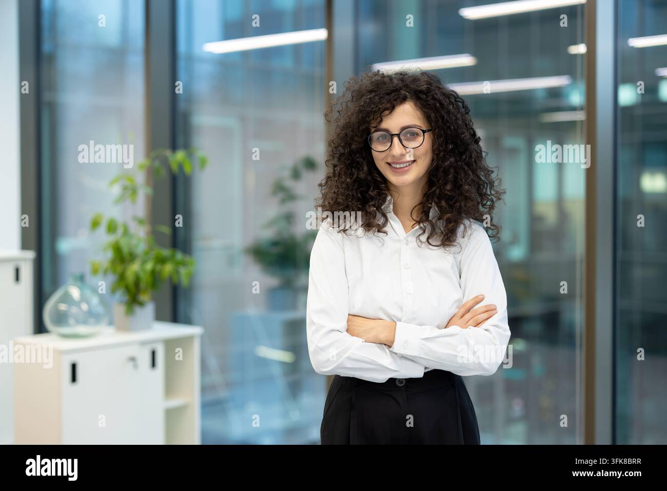 Femme d'affaires professionnelle aux cheveux bouclés et lunettes debout dans un bureau moderne. Cette employée féminine confiante sourit les bras croisés pour un portrait d'entreprise au travail. Banque D'Images