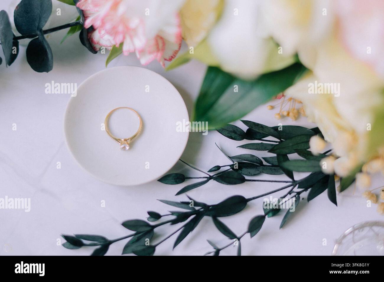 Bague de fiançailles en diamant élégante sur une petite assiette blanche avec des fleurs délicates et des feuilles vertes Banque D'Images