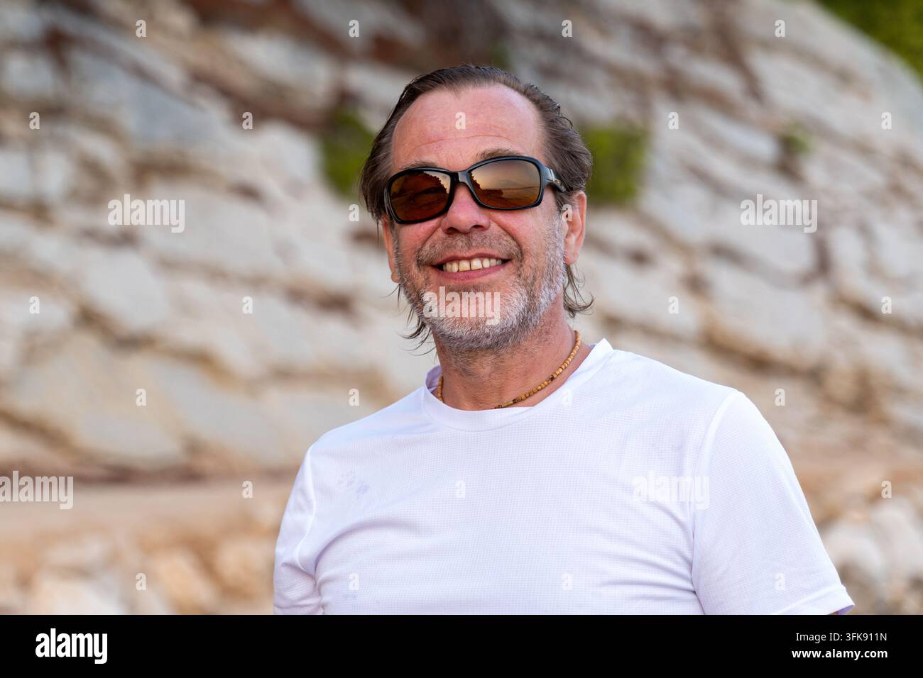 Un homme souriant d'âge moyen portant des lunettes de soleil se tient à l'extérieur devant un fond rocheux. Banque D'Images