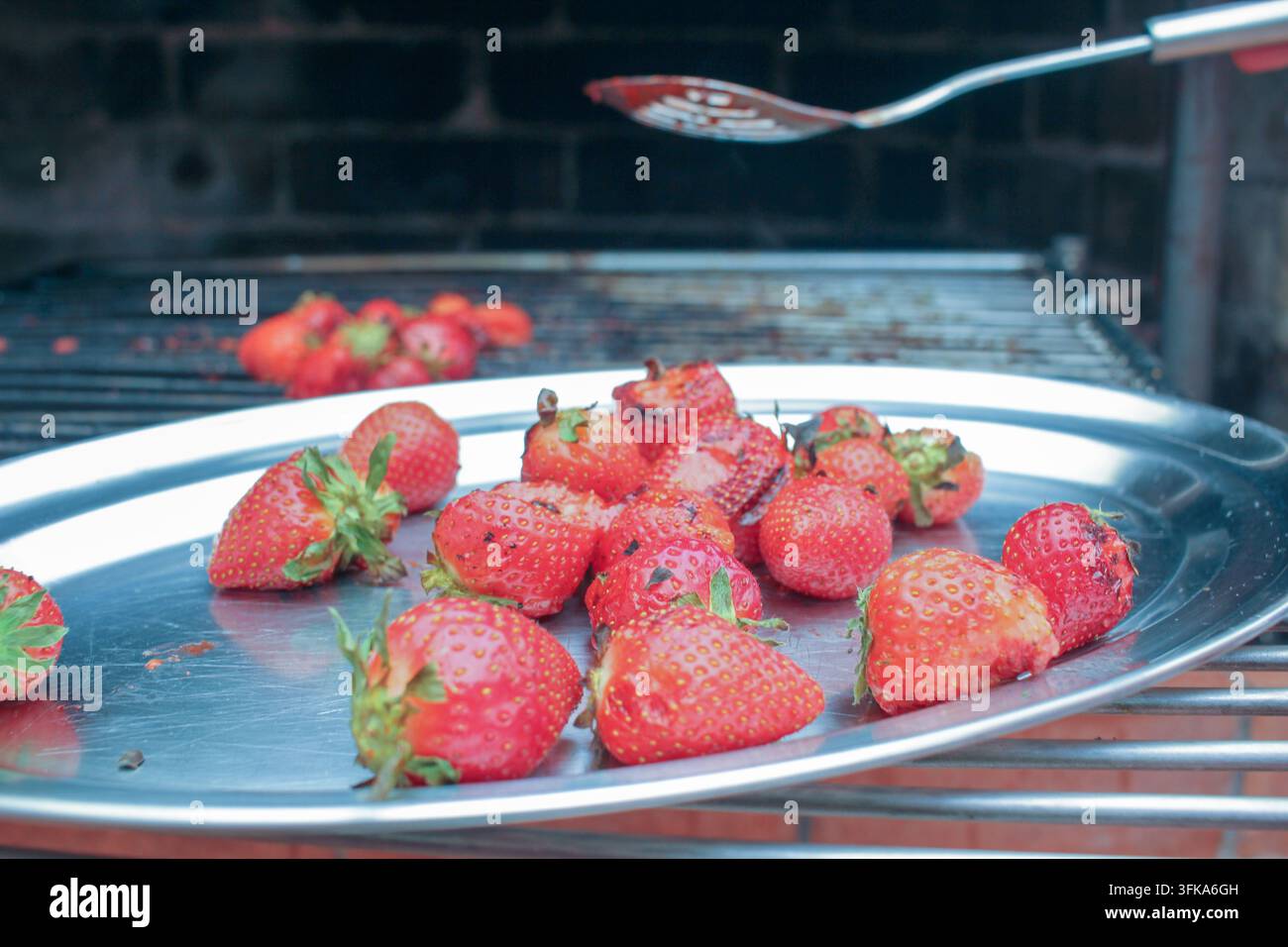 Un cliché détaillé de fraises rouges mûres disposées sur une plaque en acier avec un barbecue et une spatule en arrière-plan. Banque D'Images