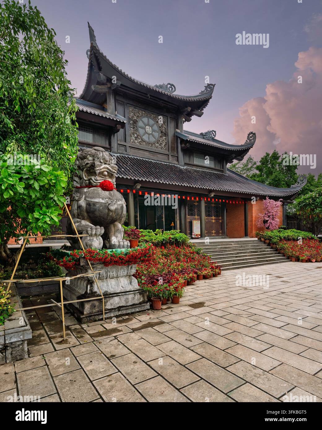 Lion de pierre à la pagode de Bai Dinh, Ninh Binh, Vietnam, présentant une architecture en bois traditionnelle et des fleurs colorées dans une cour en pierre au coucher du soleil Banque D'Images